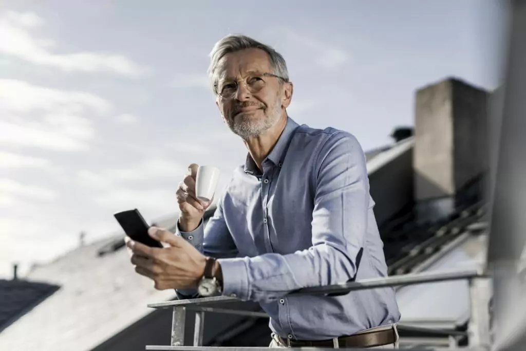 Grey-haired businessman with smartphone standing on balkony drinking coffee